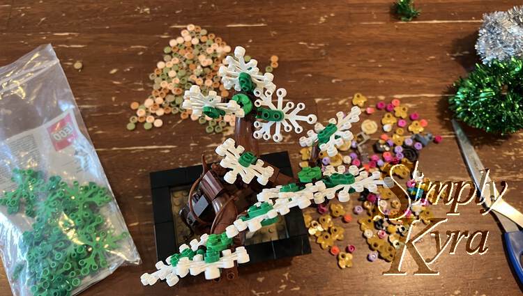 Image is taken from above looking down at the tree with green "twigs" attaching each white leaf to the brown branches. Below and around it are the green leaves in a bag, the multi-colored dirt, and a pile of the new bricks in bright colors. The tinsel and scissors are off to the right side. 