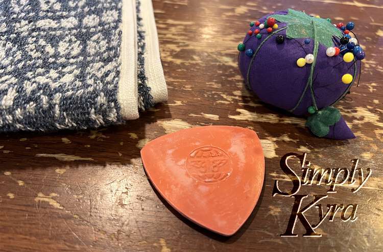 Image shows the edge of the folded towel beside a triangle of red tailor chalk and purple tomato with multi-colored sewing pins in it. 