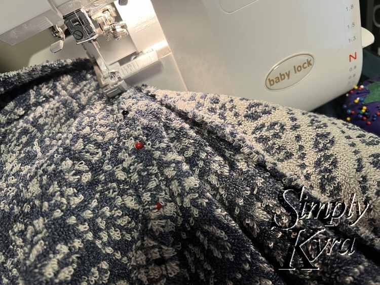 Image shows the towel half way through being cut in half with the serger. The pins are still lined up in front of the pressure foot and the cut halves are laid out behind (serged side) and to the right (unfinished side).
