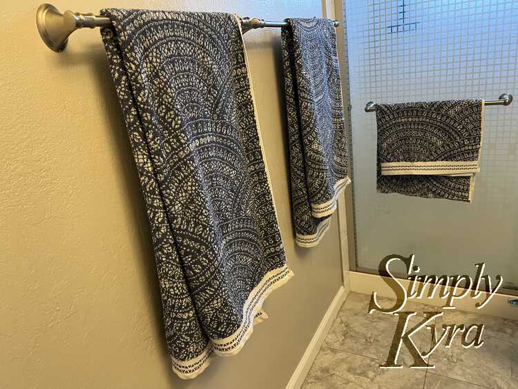 Image is taken from the side further away than the last. Here you can see two towels hung side by side and the bath mat folded and hung on the bath of the sliding shower door. 