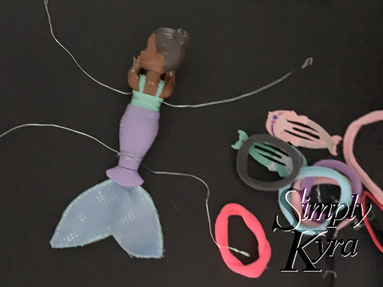 Image is taken from above showing the wire wrapped around two spots on the mermaid. To the right are the two mermaid hair clips and some colorful ponytail ties. 