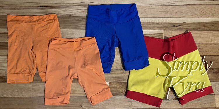 Image shows four pairs of long shorts laid out. The two on the left are orange (of different hues), then a blue one, and finally a yellow one with red bands. 