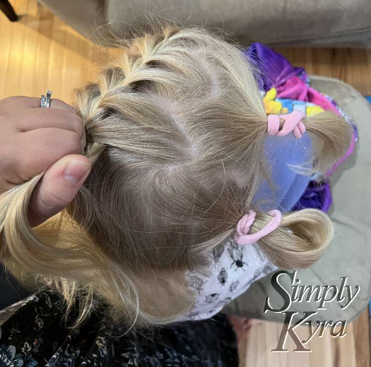 Image is taken from above looking down at Zoey's head. There's a zigzag part running down the center with the right side hair collected into two pink ponytails. The left side is French braided to the back top of her head as my left hand holds the strands in place.