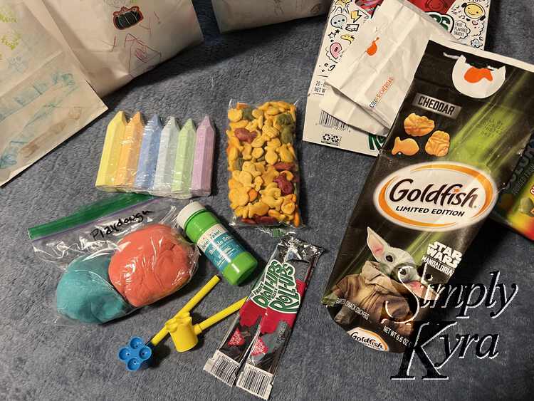 Image show the contents of a treat bag including a mix of homemade (playdough), store-bought (chalk, bubble stuff, fruit rollups), and repackaged (goldfish crackers in a snack bag). The white decorated bags are in the back and the empty packaging (fruit rollup and goldfish crackers) are to the right. 