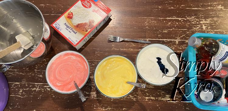 Image shows three bowls side by side. From left to right they're red, yellow, and white with blue dye in the center. Left of this is an empty and dirtied stand mix bowl with spatula and an empty Betty Crocker mix. To the right is a blue basket of food dye and flavor extracts. 