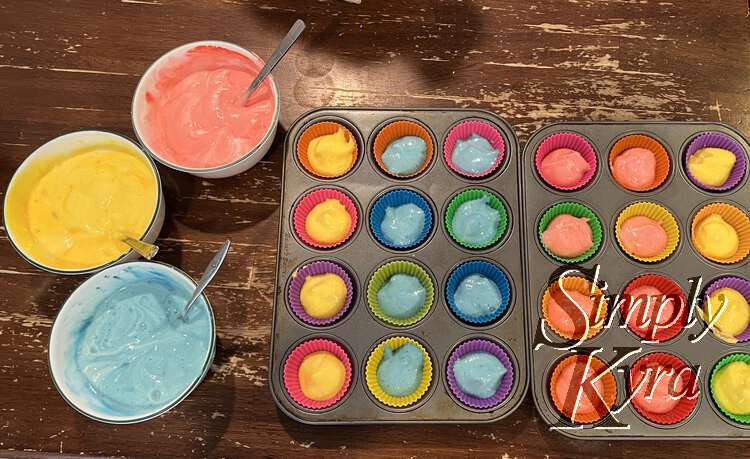 Image shows two dozen cupcakes halfway filled up. From left to right there are four yellows, eight blues, eight reds, and four more yellows. The bowls of colorful angel food cake batter are to the left with spoon handles sticking out.