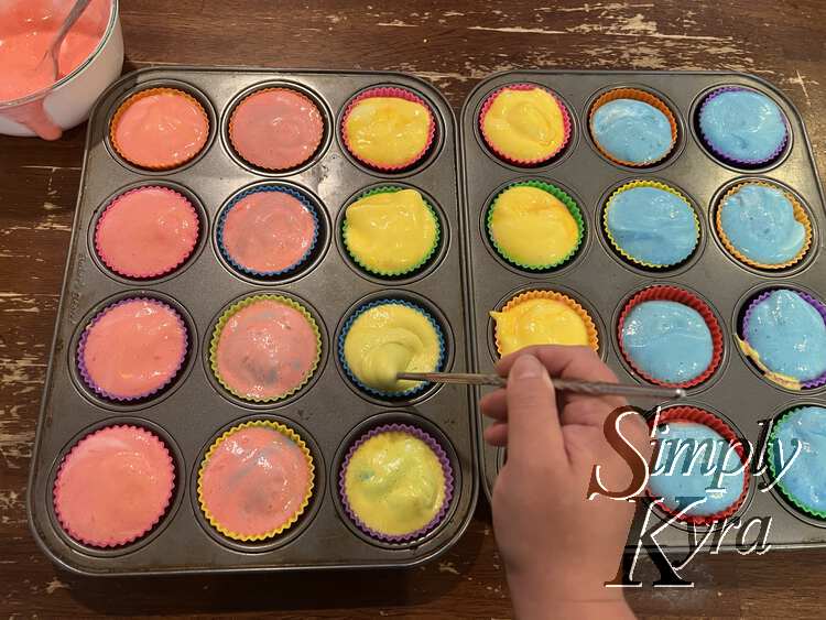 Image shows the same two cupcake tins but now the far right ones are blue. My hand is poised over a second one stirring the contents with a metal chopstick. The mixed ones show a bit of the orange, purple, or green through the marbling of the top and bottom colors.