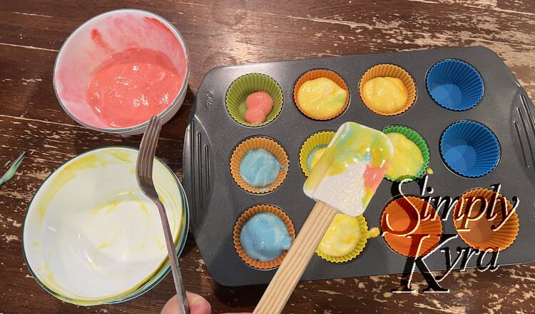 Image shows a larger muffin tin with most of the liners filled in. To the left sits the red bowl and an emptied yellow bow. In the foreground there's a fork and multicolored spatula. 