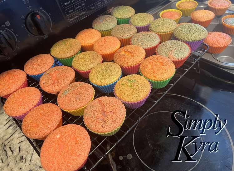 Image shows two dozen colorful cupcakes lined up on a cooling rack while the rest sit propped up between the muffin divots on the other tin.