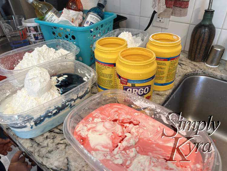 Image shows three small clear containers with cornstarch and dyed water partially mixed. In the background sits three empty Argo cornstarch containers and a container of food dyes. 