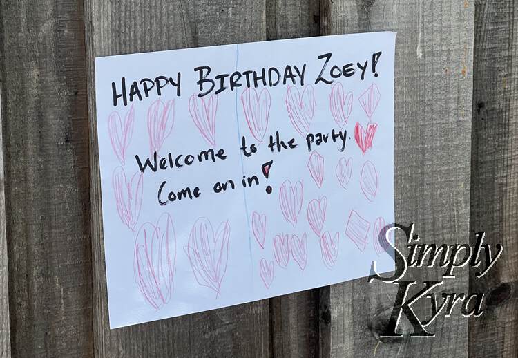 Image shows a white rectangular sign against wood saying "Happy Birthday Zoey!" "Welcome to the party. Come on in!". Surrounding the words there are pink hearts covering the page. 