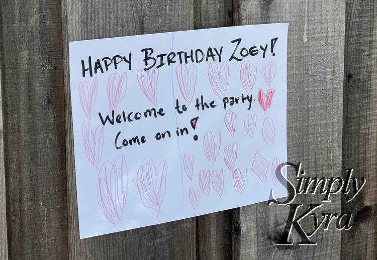 Image shows a white rectangular sign against wood saying "Happy Birthday Zoey!" "Welcome to the party. Come on in!". Surrounding the words there are pink hearts covering the page.