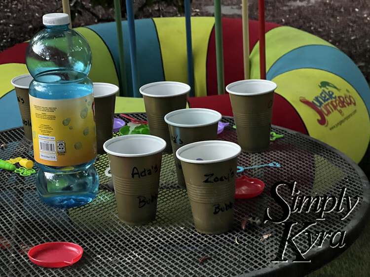 Image shows an outside table with labeled disposable cups, bubble wands and trays, and a. tall bottle of bubble mix. In the background is a large Jungle Jumparoo for ht kids to hop and swing on. 