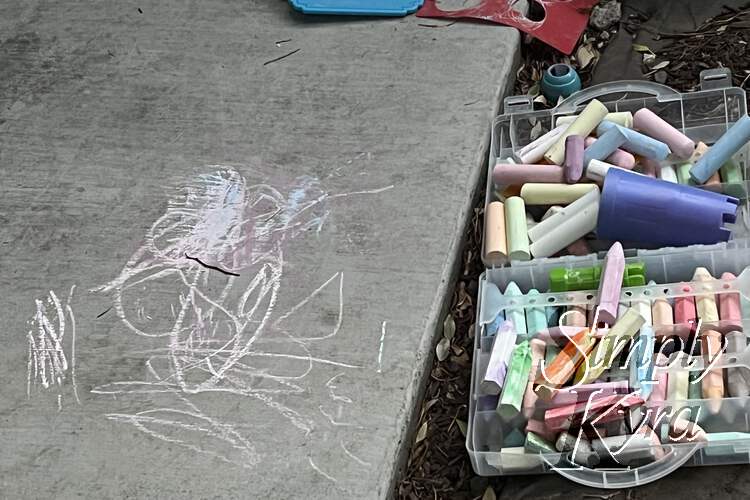 Image shows an opened plastic suitcase filled with chalk next to the sidewalk with some white and pink doodles. 