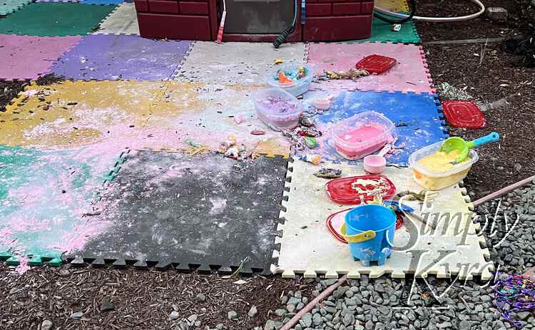 Image shows multicolored interlocking foam squares with pink oobleck spread out on it and the containers, shovels, and toys to the right. 