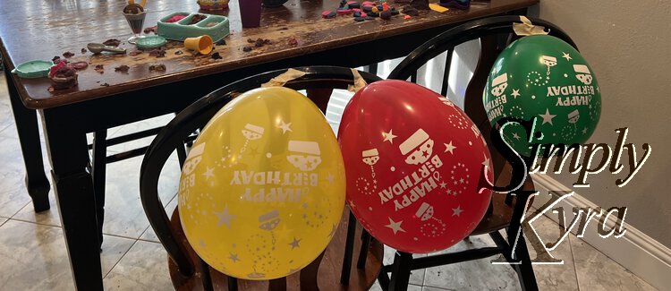 Image shows a red and yellow birthday balloon taped to the left chair and a matching green one on the right chair. In the background you can see kinetic sand and toys on the table.