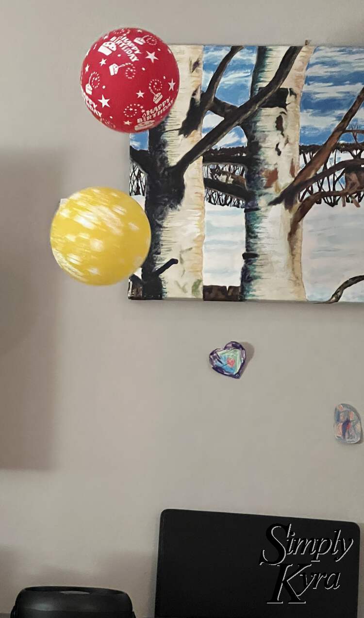 Image shows two balloons hovering in the air in front of a painting and above black fan. Both are a bit blurred although you can tell the yellow one is spinning faster as it's more blurred. 
