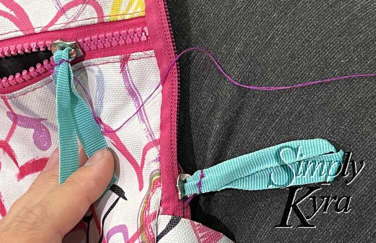 Image shows a closeup of the two teal ribboned zipper pulls. On is fully stitched together while the other one has the end of purple thread coming from it. 