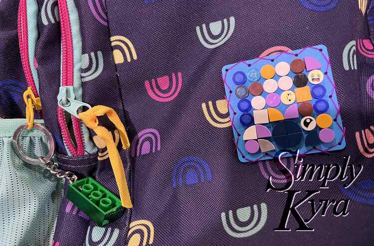 Image shows a closeup of Zoey's rainbow backpack with a green brick keychain, a yellow ribbon, and the LEGO® patch edged with purple thread and festooned with random LEGO® dot circles, squares, curves, an eye, and two emojis. Other than a black circle it looks random and helter skelter. 
