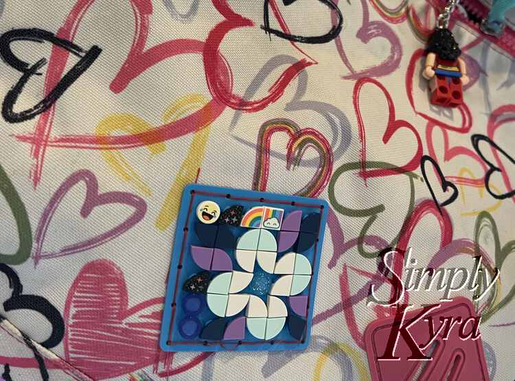 This image shows a closeup of Ada's heart backpack with a view of her patch and, in the background, the back of her Wonder Woman LEGO® keychain hanging from a zipper. 