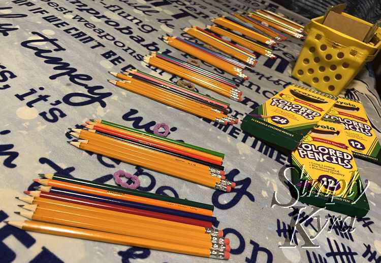Image shows the yellow basket for the school supply stash, the empty colored pencil boxes, and a row of piles of writing instruments and a small ponytail tie.