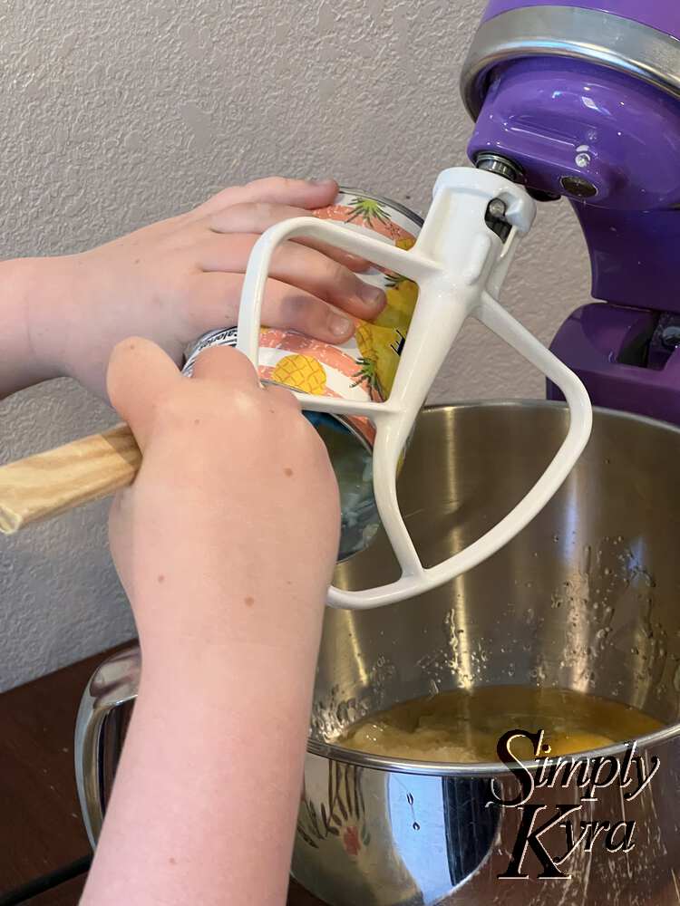 Image shows Ada using a spatula to scrape the pineapple can contents into a stand mixer bowl. Below you can see an egg sitting there already.