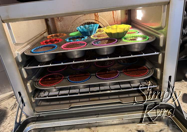 Image shows two trays of brownie in colorful liners in the small oven with two extra ones laid overtop. 