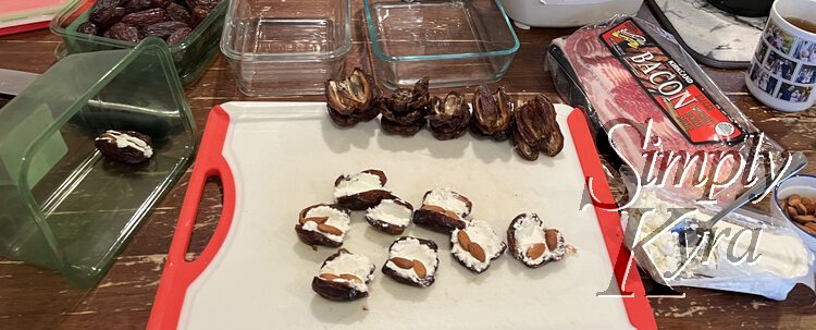 Image shows a plastic cutting board with some goat cheese and almond opened dates in the front, empty and cut open dates in the back, and closed stuffed dates in the container to the left. To the right sits the package of bacon waiting to be used and the empty glass containers waiting in the back for the finished items. 