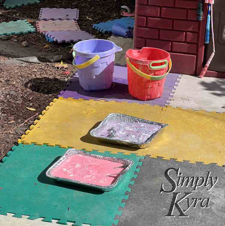 Image shows some foam squares laid out in front of a playhouse with the two (pink and purple) casserole dishes out front and two buckets. 