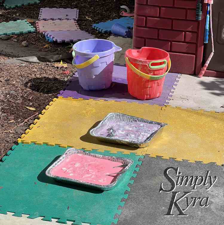 Image shows some foam squares laid out in front of a playhouse with the two (pink and purple) casserole dishes out front and two buckets.