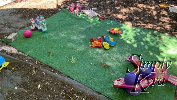 Image shows a section of fake grass with bricks holding the corners down and toys laid out carefully showcasing Ada's vision of the party. 