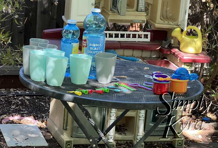 Image shows the plastic cups, two bottles of bubble mix, and wands laid out on the table with a kitchen set behind to make even more concoctions. 