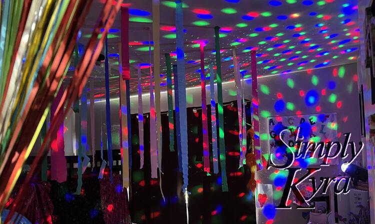 Image shows the metallic streamers pulled back like a curtain showing up the multicolor crepe streamers hanging from the ceiling and the purple, red, green, and blue lights projected everywhere. 