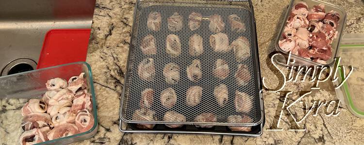 Image shows a small black cookie sheet with a grid of wrapped dates laid out. On top lays a basket tray about to be decorated. To either side sits two containers with more dates inside. 