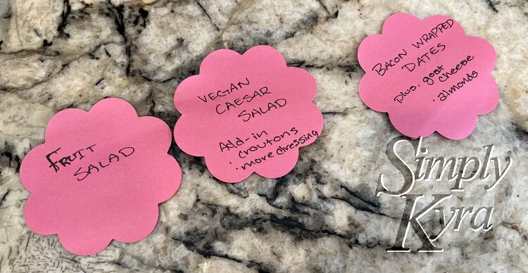 Image shows three pink flowered notes lined up on the counter with the name of the recipe, any add ins (croutons and dressing for the salad), and any potentially problematic foods (cheese and almonds on the bacon wrapped dates).