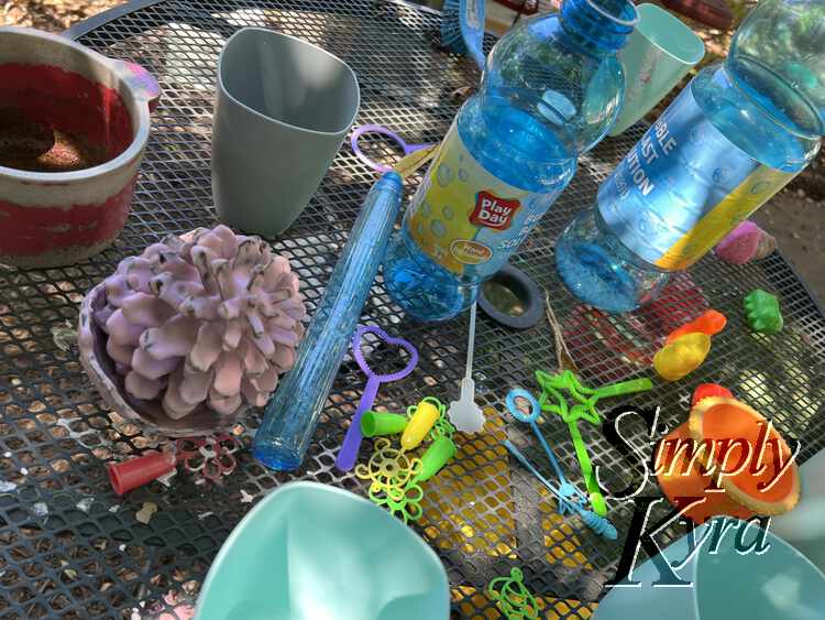 Image shows the same table as above with empty bubble mix bottles, cups, wands, kitchen set pot, and a pink oobleck coated pinecone. 