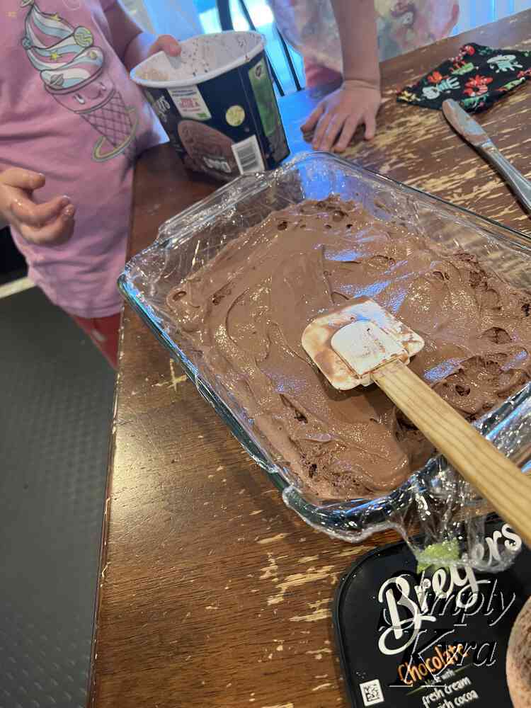 Image shows the the mostly smoothed out chocolate ice cream in the plastic wrap lined glass casserole dish with a spatula over top. The Breyers container lid is in the front of the photo while the container itsel is behind surrounded by either kid.
