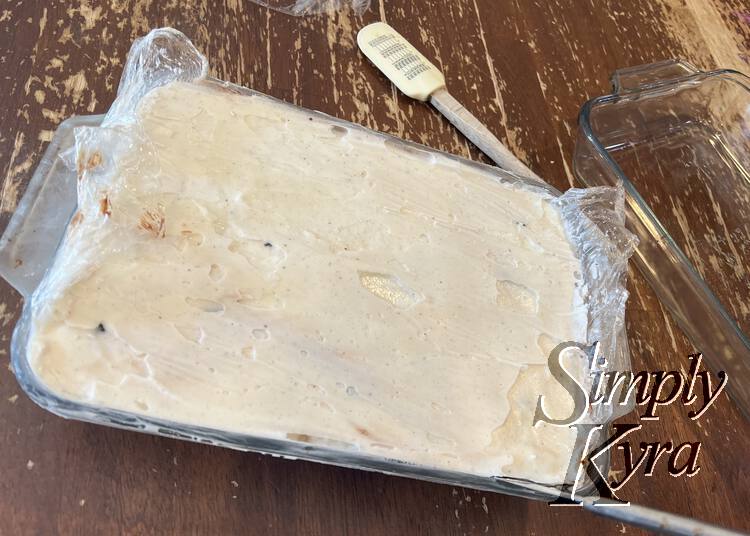 Image shows the smoothed out cake with plastic wrap sticking out the sides and a spatula and another glass dish in the background.
