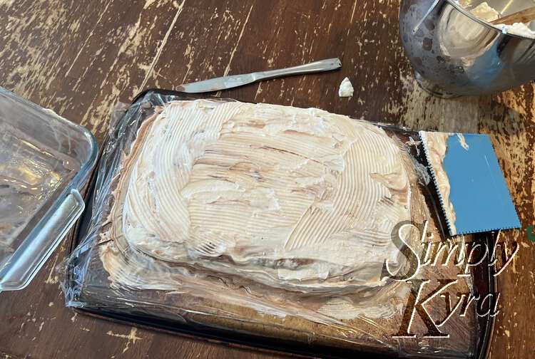 Image shows a line streaked whip cream covered cake laid out on the underside of a cookie sheet with plastic wrap on it. 