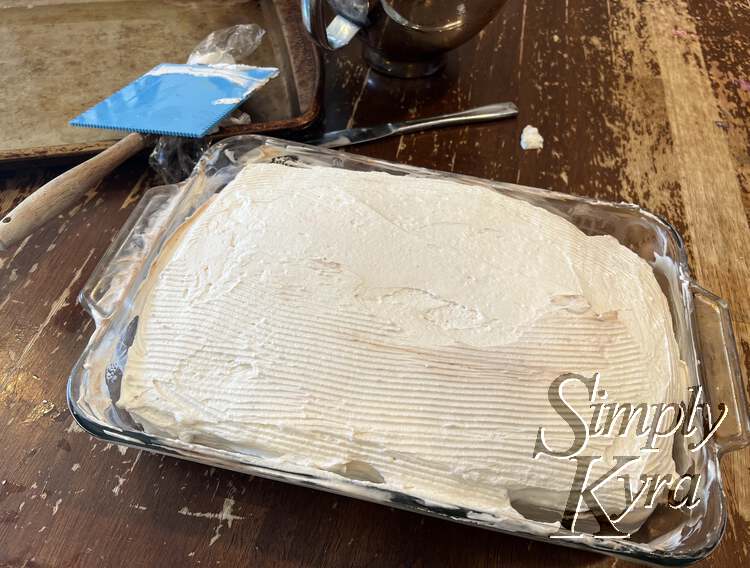 Image shows the curved cake filling the casserole pan a bit better with lines coating most of it and the top smooth-ish. The cookie sheet, plastic wrap, edging tool, and knife lay behind it. 