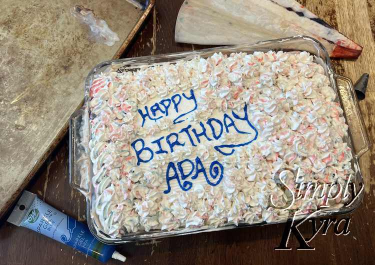 Another close photo of the cake with the entire cake coated in white, blue, pink, and purple dappled swirled flowers with an opening for the text saying "Happy Birthday Ada". In the background you can see the tube of blue gel icing, the cookie sheet, the dyed piping bag, and the tip. 