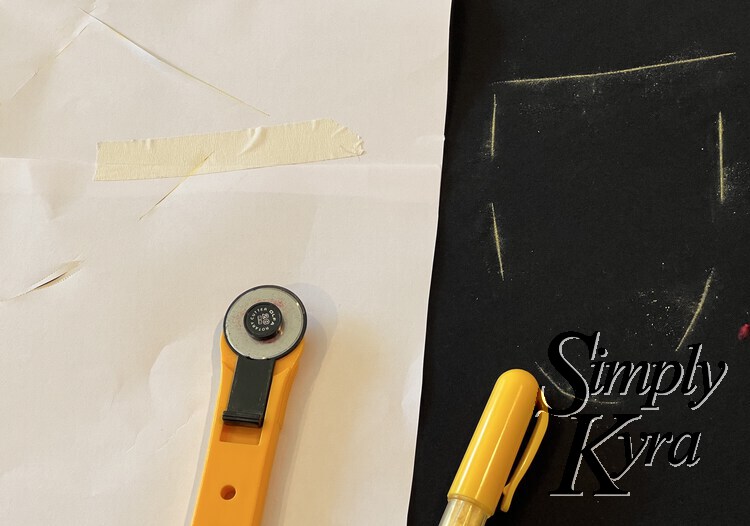 Image shows the wrong side of the paper pattern laid out offset on the black fabric. The yellow chalk marking showing the pocket outline are obvious on the black fabric. The rotary blade and yellow chalk pen are both laid out at the bottom of the image.