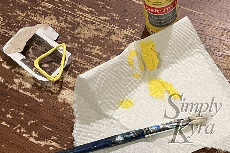 Image shows the yellow triangle laid out on a scrap of packaging. The yellow acrylic paint bottle sits behind a yellow smudged paper towel and a paintbrush. 