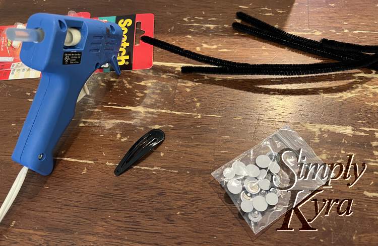 Image shows a small zipped bag of googly eyes, a simple black hair clip, a heating hot glue gun, black pipecleaners, and some emergency just in case crazy glue.