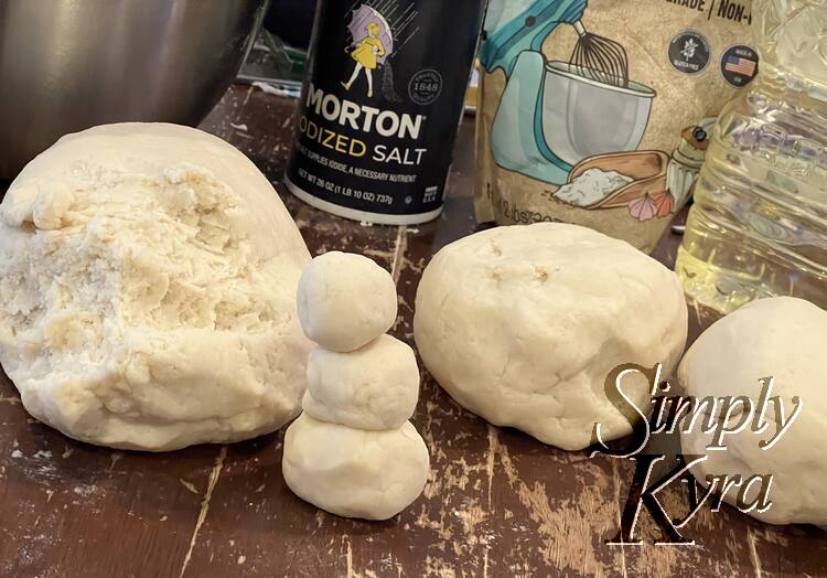 Image shows two smaller balls of play dough next to one larger one with a piece removed. Between them sits a single undecorated snowman. In the background is some salt, cream of tartar, and canola oil. 
