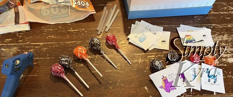 Image shows five Tootsie Roll lollipops laid out on the table. My glue gun sits to the left, the bag and extra sticks behind it, and the cards with and without the lollipop attached to the right. 