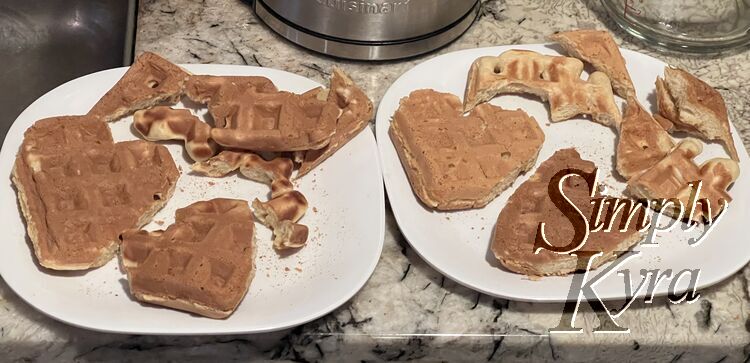 Image shows two white plastic plates side by side next to the kitchen sink beside the kettle. On each are two heart shaped waffles and, to the side, the cut off edging.