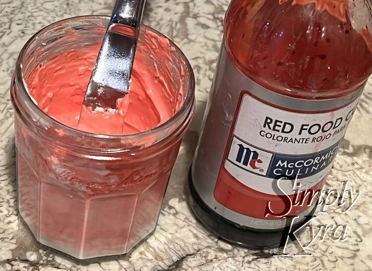 Image is a closeup, taken slightly from above, of the jar of pink cream cheese with a knife sticking out of it. Beside it is a large bottle of red food dye coloring.