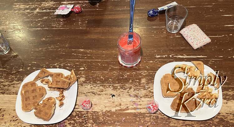 Image shows the two plates from before out on the table spaced further apart. They both contain two heart waffles and the extra pieces. Beside them on the inner side sits the small container of sprinkles while between them sits the pink dyed and softened cream cheese with a knife in it for dishing. 