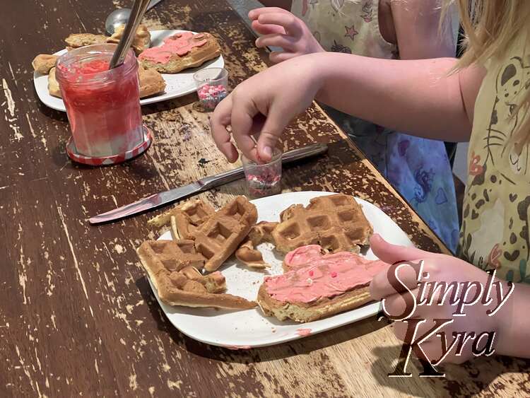 Image shows the two plates at a different vantage point so one is in the foreground and the other angled back into the background. Both have one waffle heart now pink with some sprinkles being added to it.